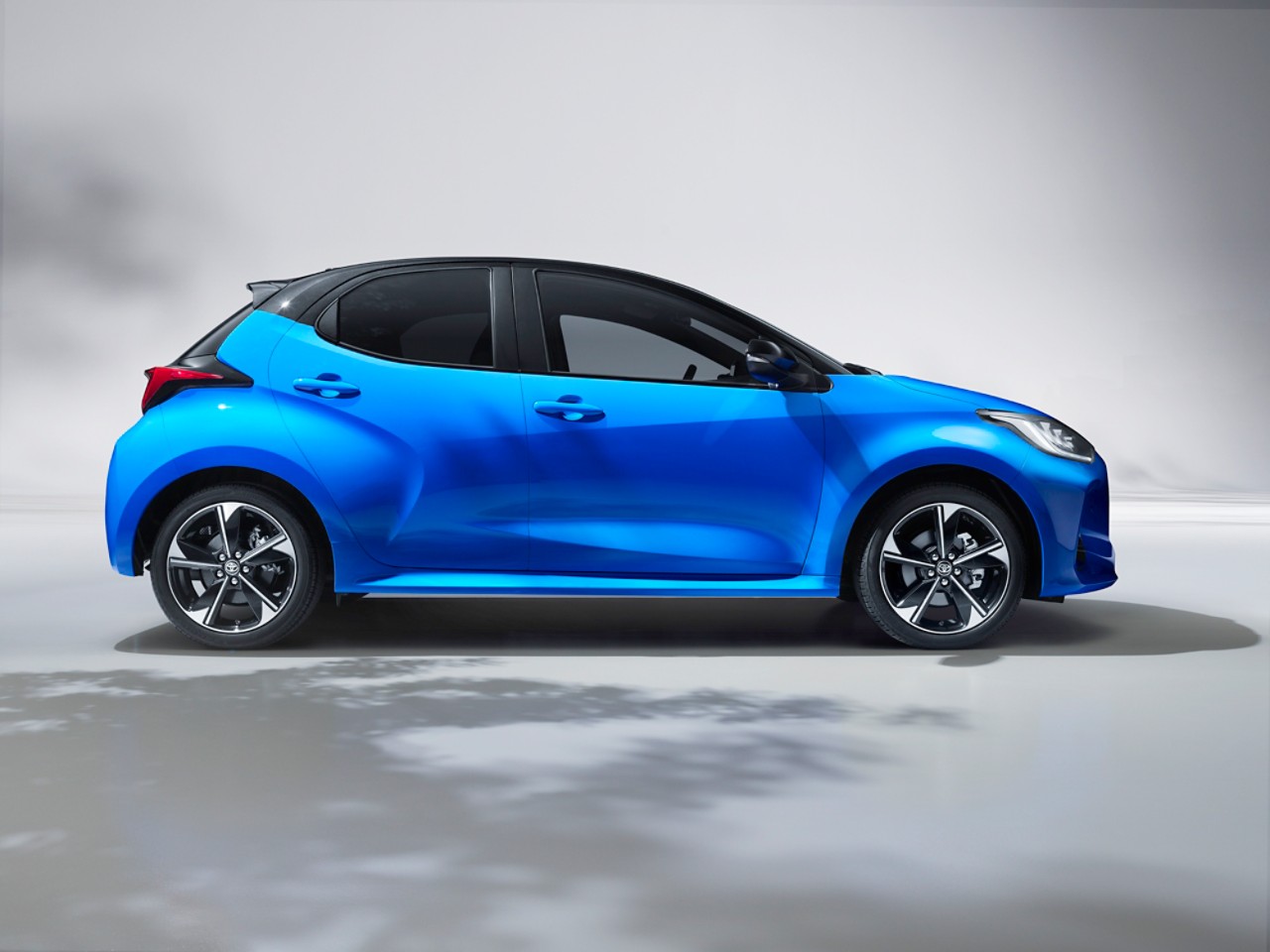 A metallic blue Toyota Yaris Hybrid car is parked in a courtyard locaOon. The shadow from a tree parOally covers the concrete floor.